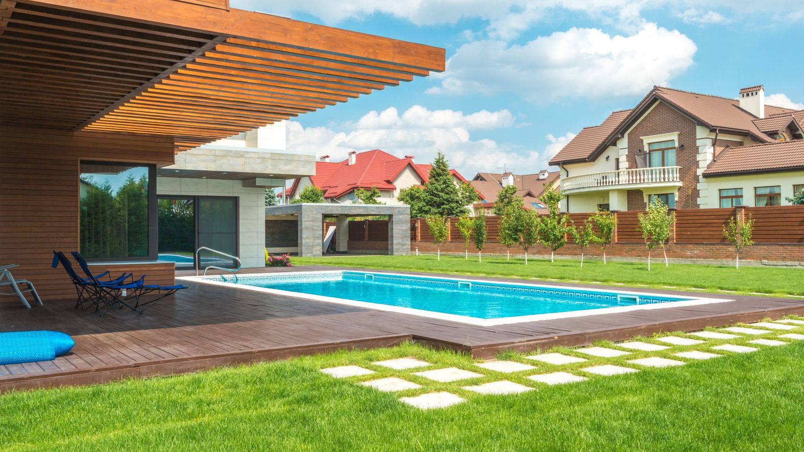 Rechteckiger Pool mit Holzterrasse im Garten eines modernen Wohnhauses, dahinter Rasen und Hecke.