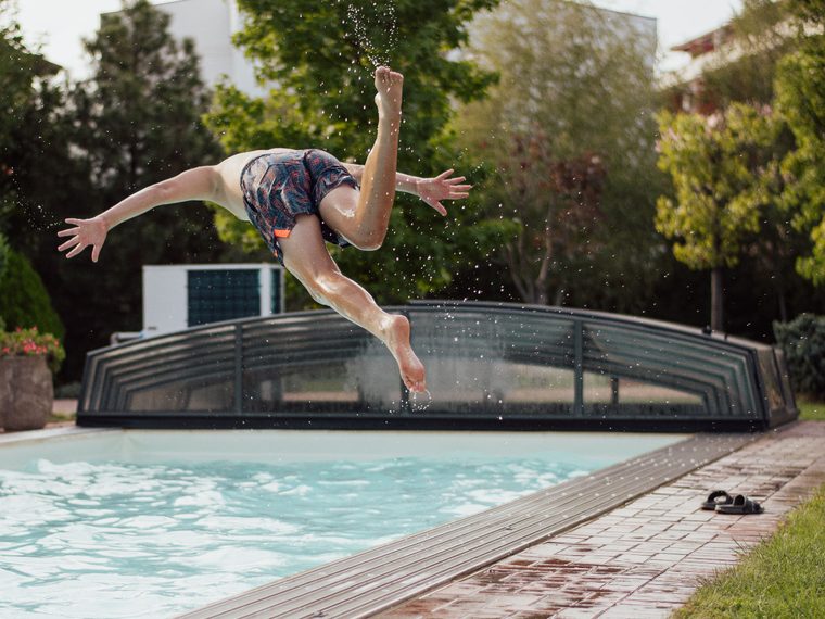 Rechteckiger Pool mit Terrasse und &Uuml;berdachung in einem Garten, eine Person springt ins Wasser.